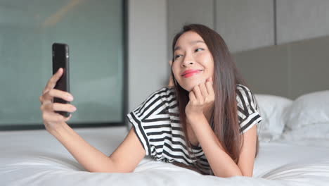 beautiful asian young smiling woman laying in bed taking selfies portraits of herself with smartphone