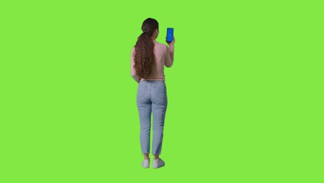 estudio de longitud completa vista trasera de una mujer sosteniendo un teléfono móvil de pantalla azul contra una pantalla verde