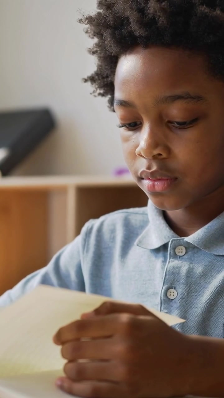 Vertical video: Starting holding book open, boy in blue polo flipping pages at home desk, studying