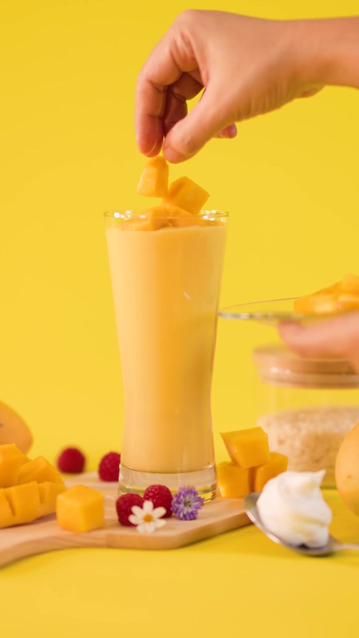 Primo piano di una mano che aggiunge cubetti di mango a un alto bicchiere da frullato su uno sfondo giallo vivace.