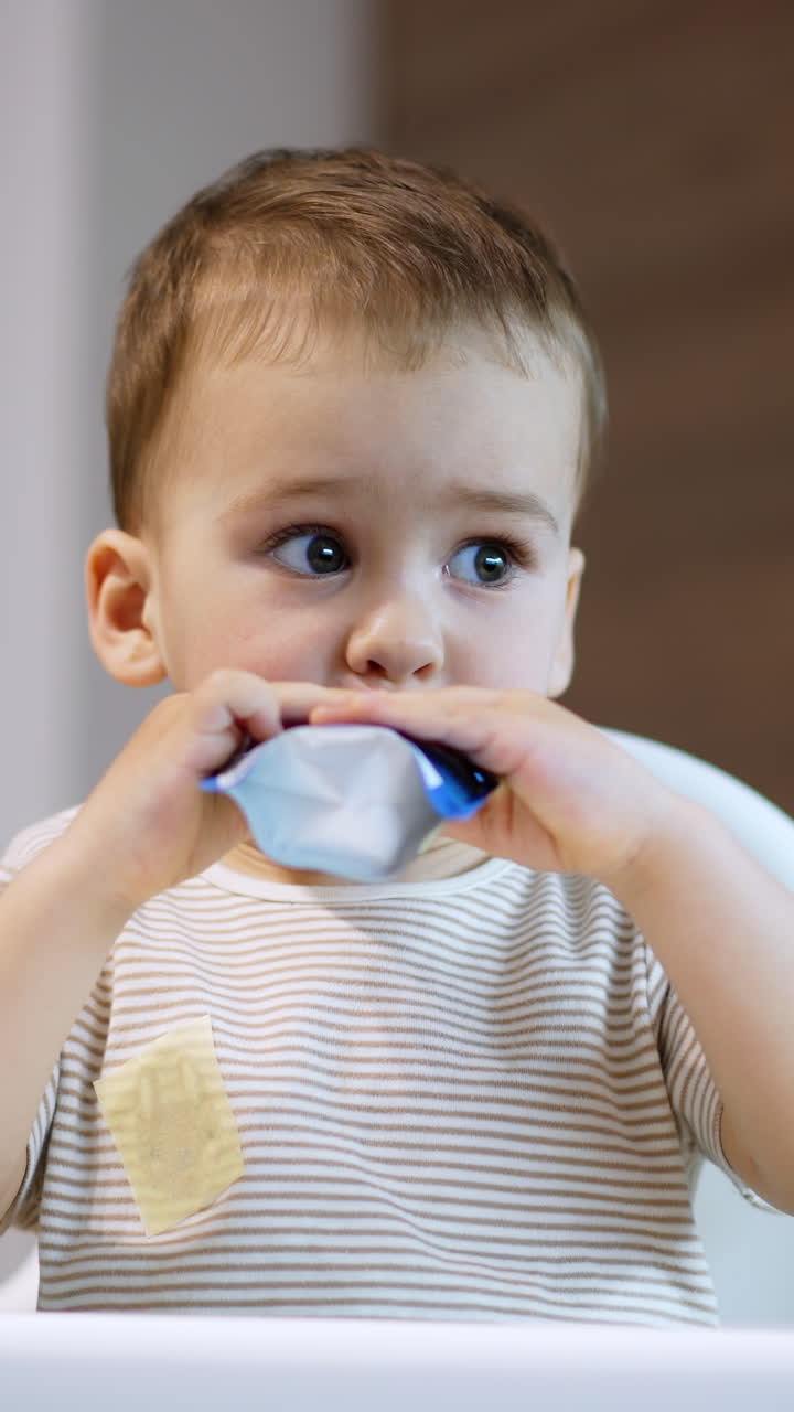 Adorable baby boy takes a pack of food into mouth. Little kid eating the pureed fruit willingly. Blurred backdrop. Vertical video