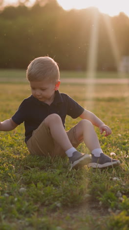 funny little boy kicks legs sitting on lush grass on playground at sunset slow motion. toddler child rests after games in park. happy childhood