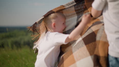 a young boy in a white shirt playfully hits a flowing plaid scarf in a grassy field. a woman and another boy are in the background, adding to the joyful and carefree atmosphere