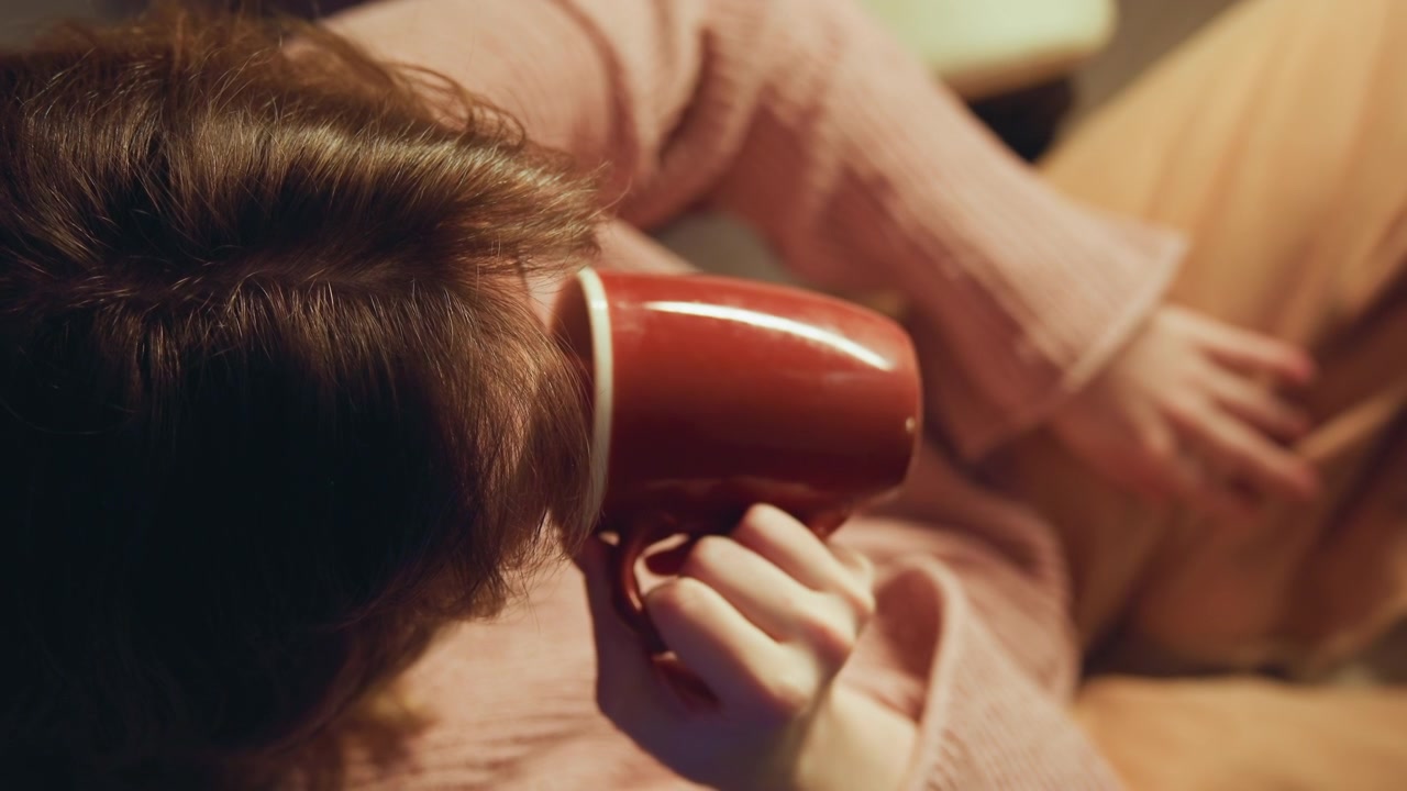 Primo piano obliquo dall'alto di una persona seduta che solleva una tazza di ceramica rossa, con il polso alzato e le dita avvolte attorno al manico, creando una sensazione di comfort, una tranquilla routine mattutina e un calore domestico silenzioso.