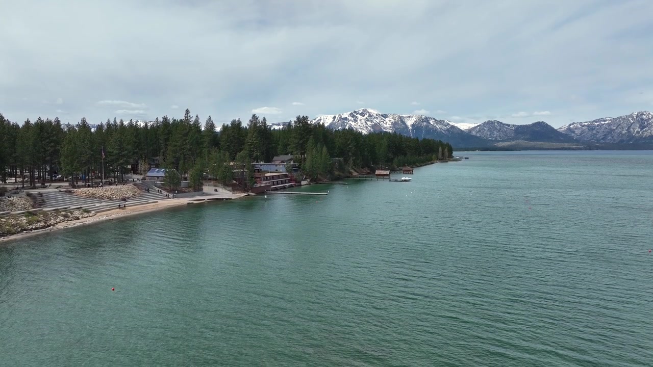 Costa panoramica a South Lake Tahoe con acqua cristallina, baite nel bosco e montagne della Sierra Nevada coperte di neve. Paesaggio alpino tranquillo e atmosfera da meta di viaggio. Vista aerea in discesa