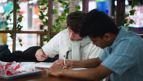 serious minded friends seated side by side sketching quietly on paper while backpacks and bags placed nearby creating hushed atmosphere with soft ambient light capturing focused creative art moment