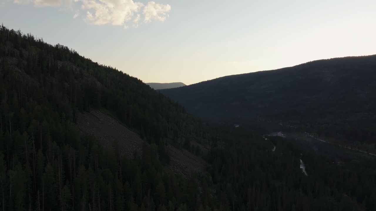 Drone right trucking shot of a valley in Uinta-Wasatch-Cache National Forest, Utah, at sunset with the Provo River surrounded by pine trees, Highway 150 to the right, mountains and a clear summer sky