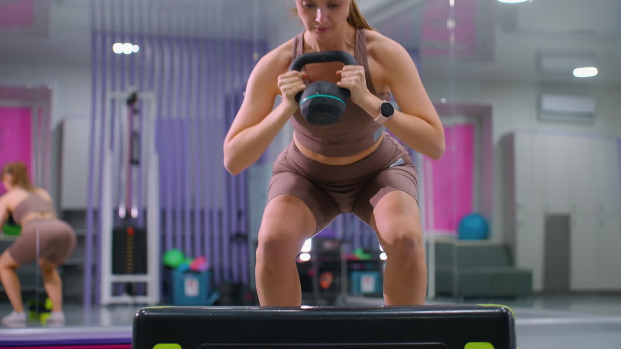 kettlebell을 들어 올리는 동안 높은 표면에 발을 고 운동을 반복하는 젊은 여성, 운동 장비를 가진 체육관 환경