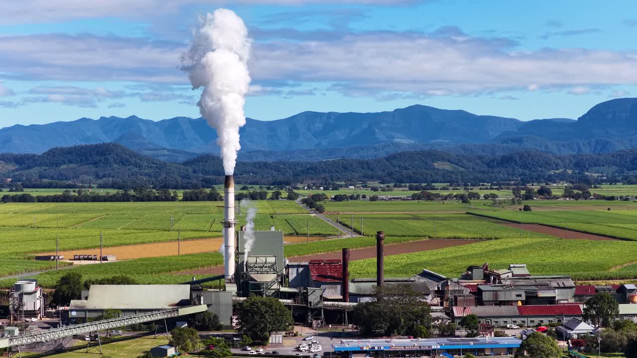 Aerial footage of a sugarcane factory emitting smoke, surrounded by lush fields and mountains under clear skies