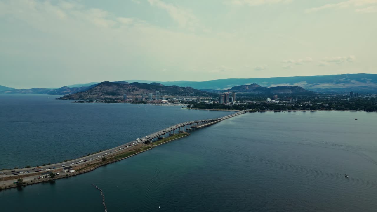 High flying Aerial over Okanagan Lake view of William R Bennett Bridge cars trucks traffic commuting Kelowna BC over calm blue water mountainous background light blue sky wispy clouds
