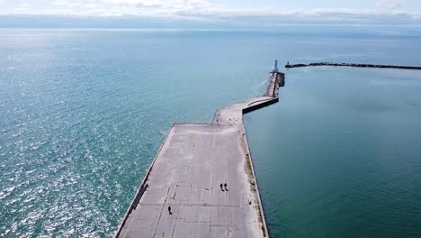 people walking on pier toward lighthouse and marina breakwater in turquoise blue lake water