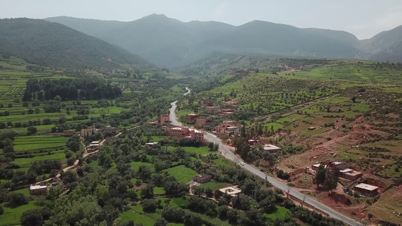 Ampia ripresa aerea di una strada asfaltata che serpeggia attraverso una verde valle tra le montagne dell'Alto Atlante. Il filmato mostra l'agricoltura terrazzata, case rurali sparse e il paesaggio pittoresco del Marocco, N