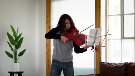 latino musician with long hair in jeans and grey shirt playing song from sheet music on red viola in white studio