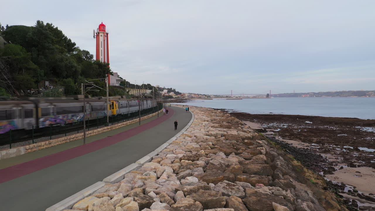 Aerial shot over People riverside walkway with trains crossing by and red Lighthouse, Lisbon