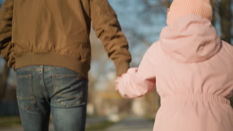 close-up of a joyful father in a brown jacket and black jeans walking and playfully jumping with his daughter in a pink cap and jacket in the park