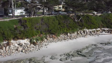 Toma-Aérea-De-Una-Playa-árida-Y-Rocosa-Junto-Al-Mar-Con-Un-Acantilado-Y-Casas-Al-Fondo