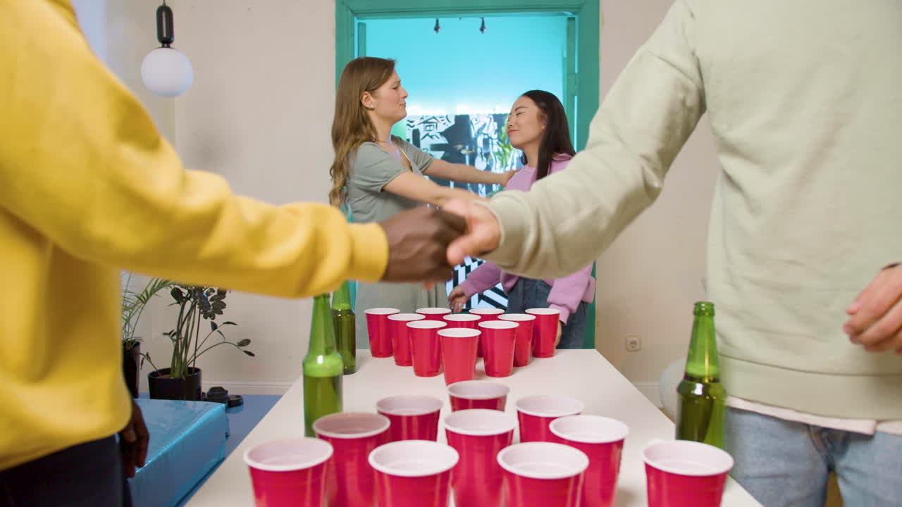 Multiethnic young friends playing beer pong at home. Asian girl throwing the ball, missing the shot and hugging her teammate while the opposing team shaking hands and giving a gist bump.