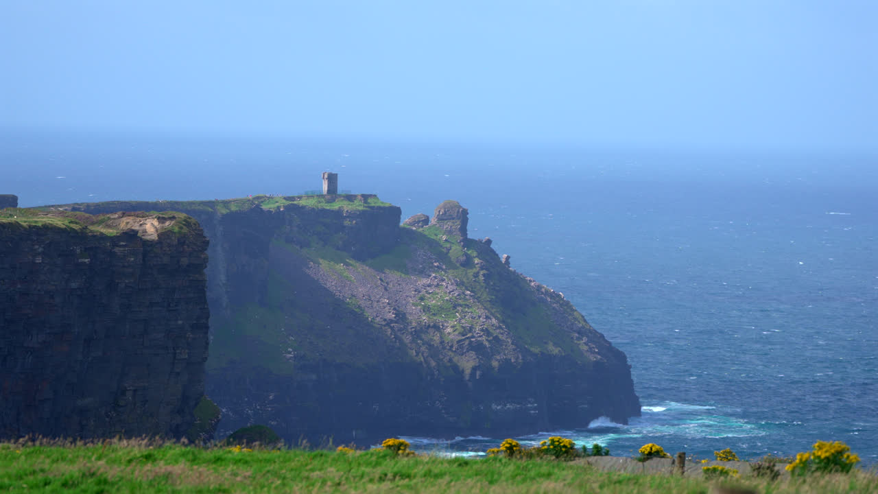 The Cliffs of Moher dominate the coastline, with a solitary tower visible in the distance. The contrast between steep rock faces and the open ocean highlights Ireland’s dramatic landscape.