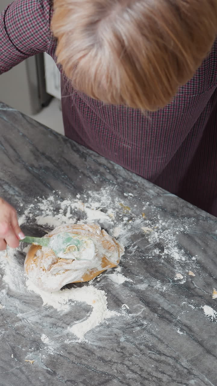 Overhead view of baker kneading soft dough on floured tiled table while resting left hand, kitchen workspace covered in flour as homemade baking process unfolds