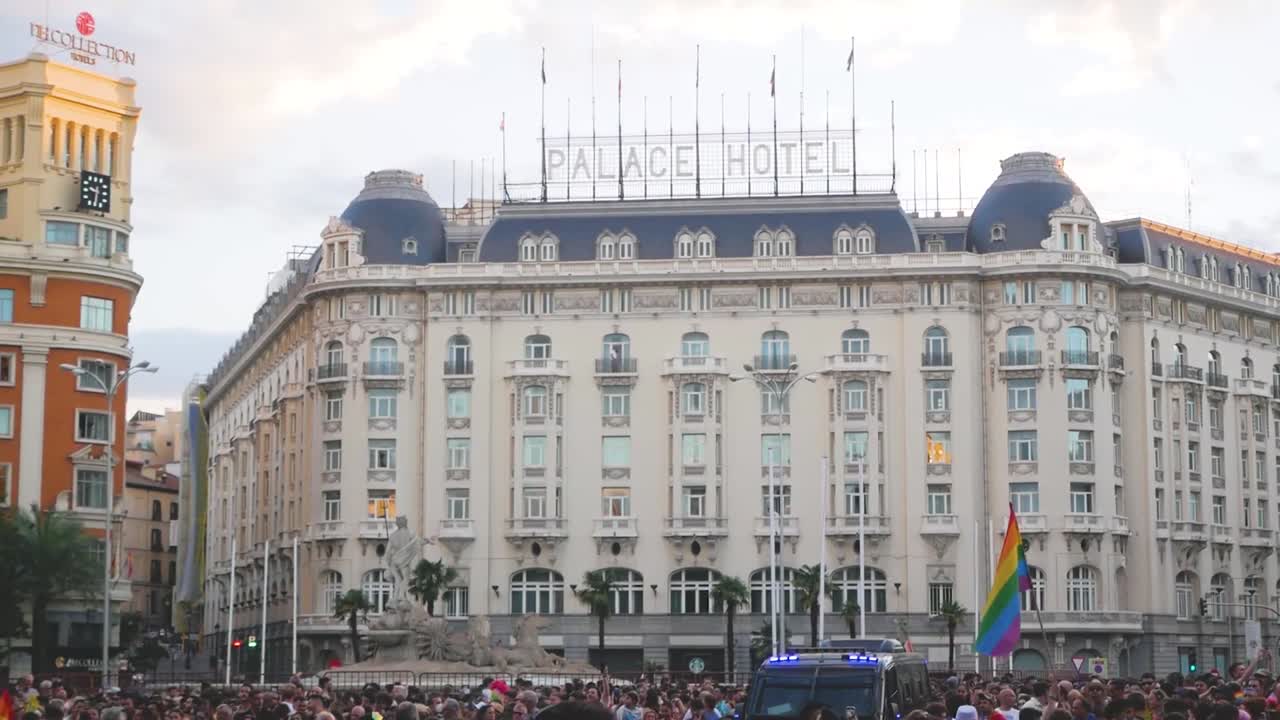 A vibrant pride protest outside the Palace Hotel in Madrid, featuring colourful banners and enthusiastic LGBTQ+ supporters.