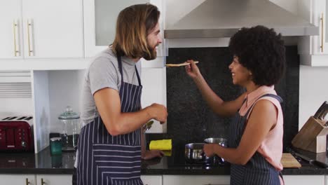 pareja de raza mixta usando delantales probando comida juntos en la cocina
