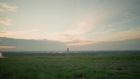 an adult male wearing a white shirt, hat, and jeans stands contemplatively in a grassy field by a tranquil lake at sunset. he gazes into the distance, reflecting on the serene landscape