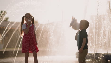 couple of little siblings eats candy floss near fountain