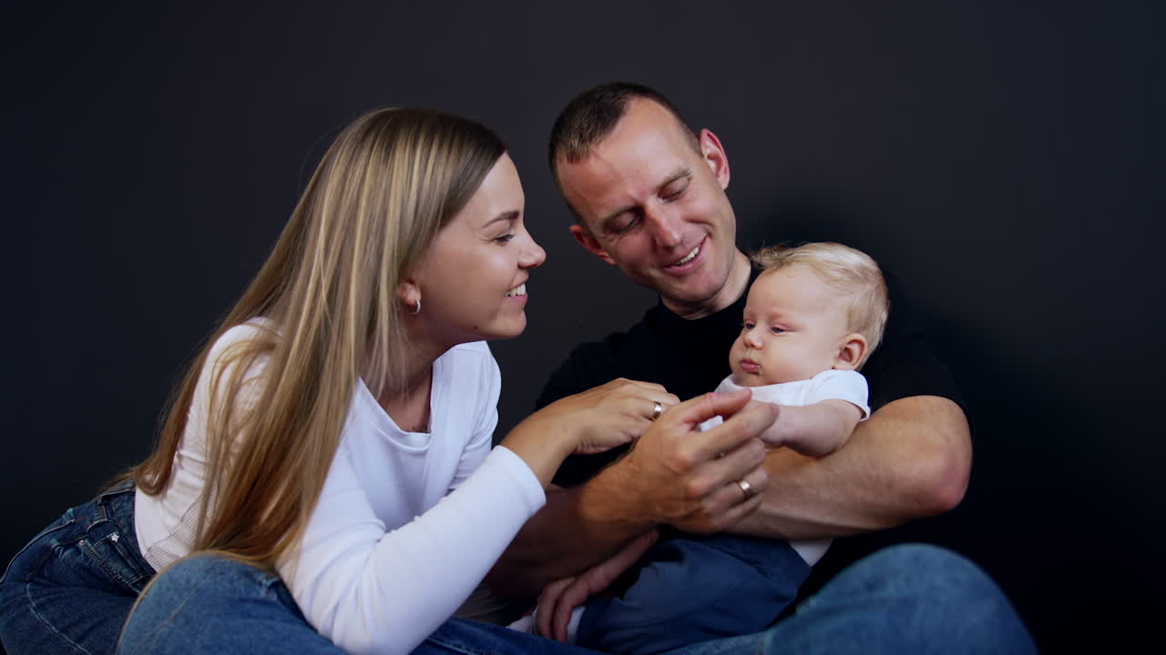 Father holds his tiny serious baby boy. Dad waves kid's hands and mother talks and smiles to her son.