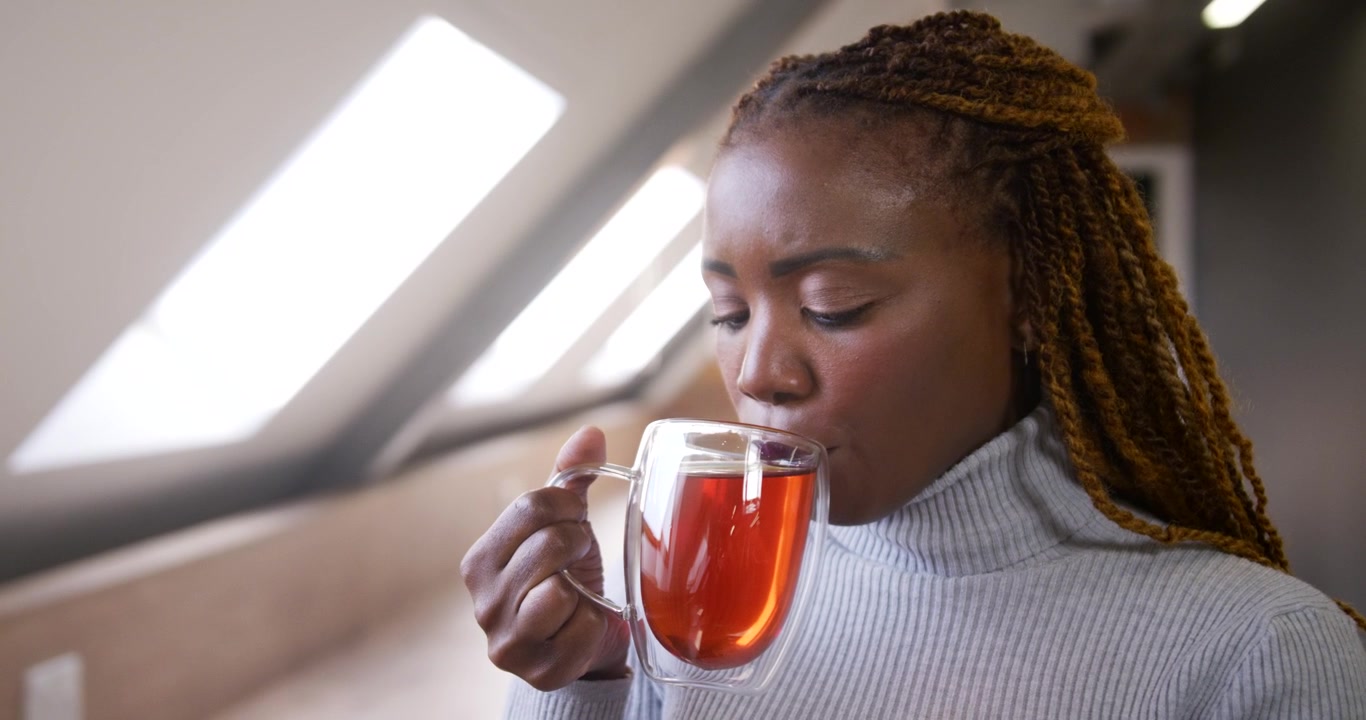 Mulher africana levantando uma caneca transparente, cheirando e tomando um gole de chá, saboreando a calma sob as claraboias do loft.