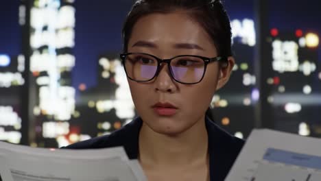 a woman with glasses is focused on analyzing documents in a modern office surrounded by city lights. the atmosphere reflects a late-night work environment working on critical tasks.