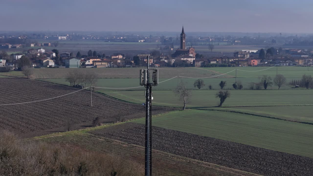 Drone orbits 5G antenna in Folgarole countryside—surrounded by green fields, power lines, and distant village skyline with church steeple in Piacenza, Emilia-Romagna, Italy