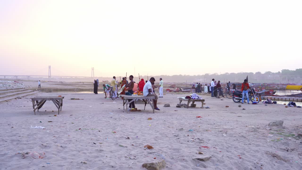 People on a sandy riverbank with boats and a bridge in the distance