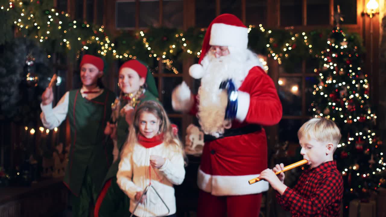 Santa Claus is dancing with children who play musical instruments in the house with a Christmas tree