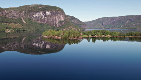 Lenta-Toma-Aérea-Desde-Una-Plataforma-Rodante-Empujando-Una-Pequeña-Isla-En-Byglandsfjord-En-Noruega-En-Un-Día-Soleado,-Los-Acantilados-Cubiertos-De-árboles-Se-Reflejan-Perfectamente-En-Las-Tranquilas-Aguas-Del-Lago