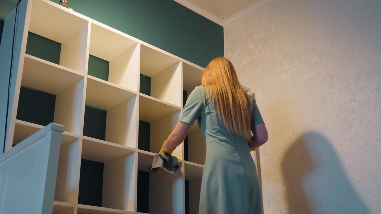 Donna che pulisce gli scaffali di casa con libri, indossando guanti gialli e usando un panno, che sistema gli oggetti decorativi in uno scaffale a cubi bianco contro una parete color ottanio, compito domestico metodico e concentrato e routine ordinata.
