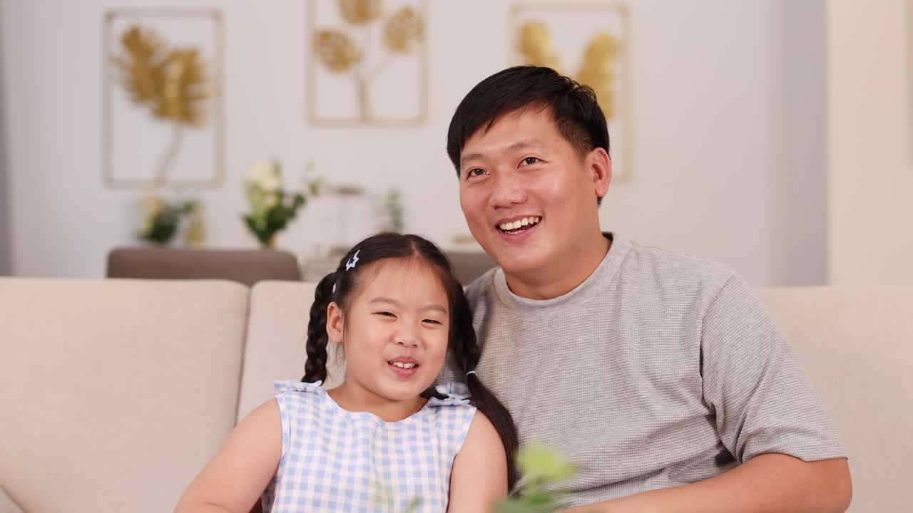 A father and daughter enjoy a playful, happy moment on a cozy couch in a warmly lit living room