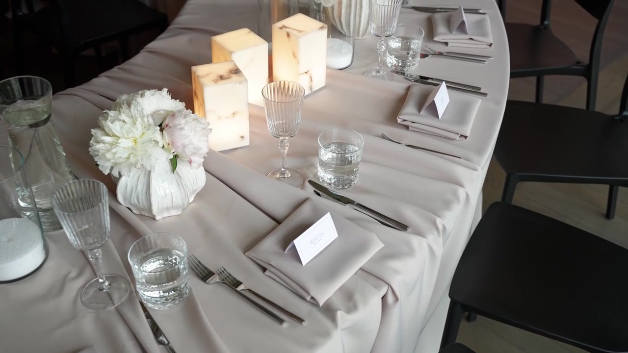 Beautifully arranged dinner table with white flowers, glowing pillar candles, crystal glassware, and minimalist place cards in soft natural light—perfect stock footage for events, weddings.
