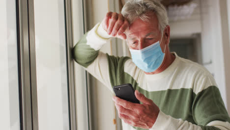 Hombre-Caucásico-Mayor-Con-Mascarilla-Usando-Un-Teléfono-Inteligente-Cerca-De-La-Ventana-En-Casa