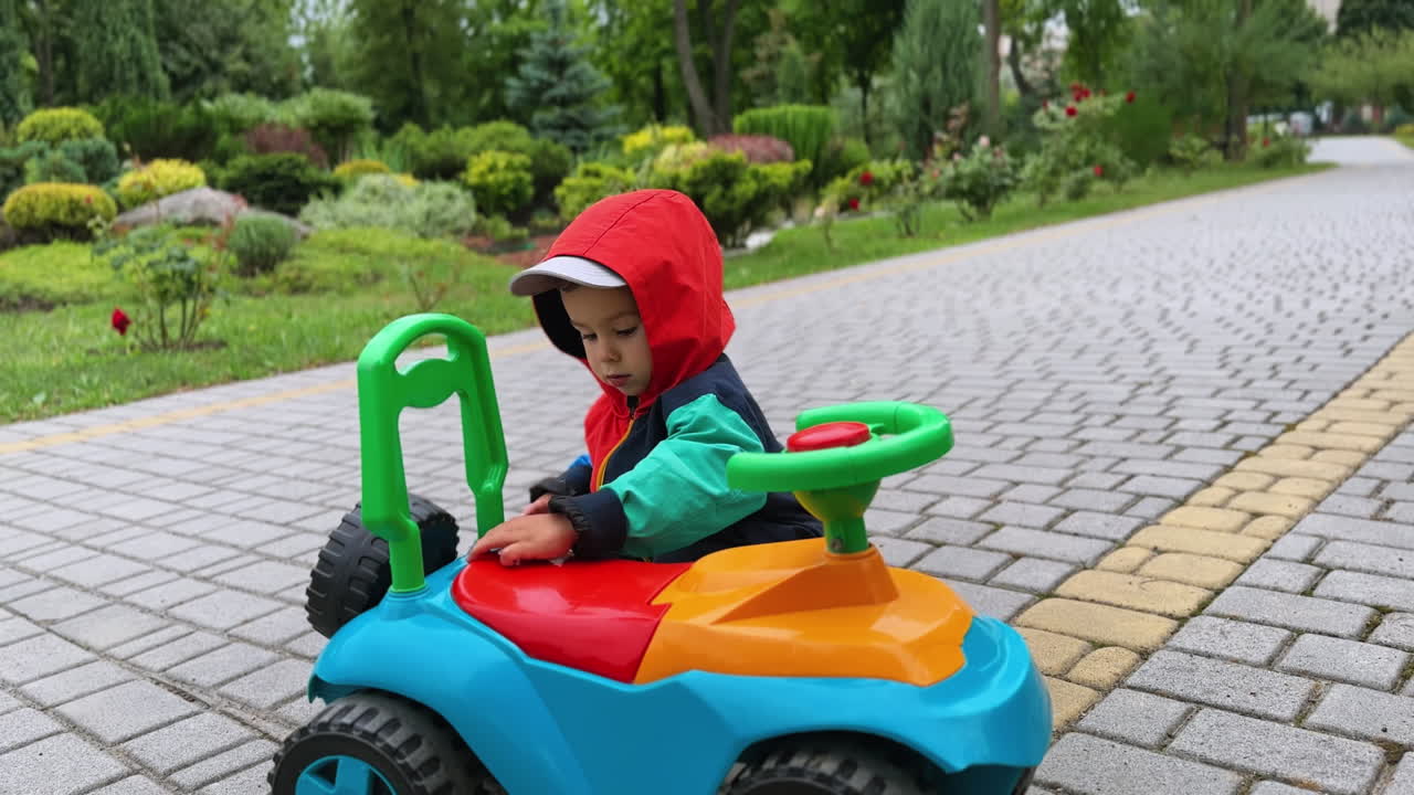 Lovely cute baby boy in jacket and hood stands near his toy car. Kid touches parts of car in the park.
