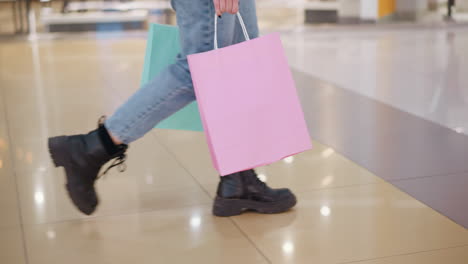 vista inferior de la pierna de una mujer elegante con botas negras y vaqueros, cargando bolsas de compras rosas y menta mientras camina junto a una tienda en un centro comercial concurrido, otros compradores que pasan añaden movimiento a la escena