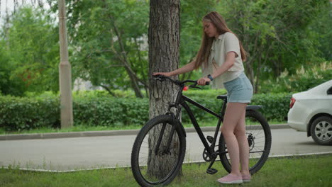 woman in jean shorts approaches bicycle leaning against tree, removes stand, and begins pushing bike in park setting with lush green background of trees, paved path, and nearby parked vehicle