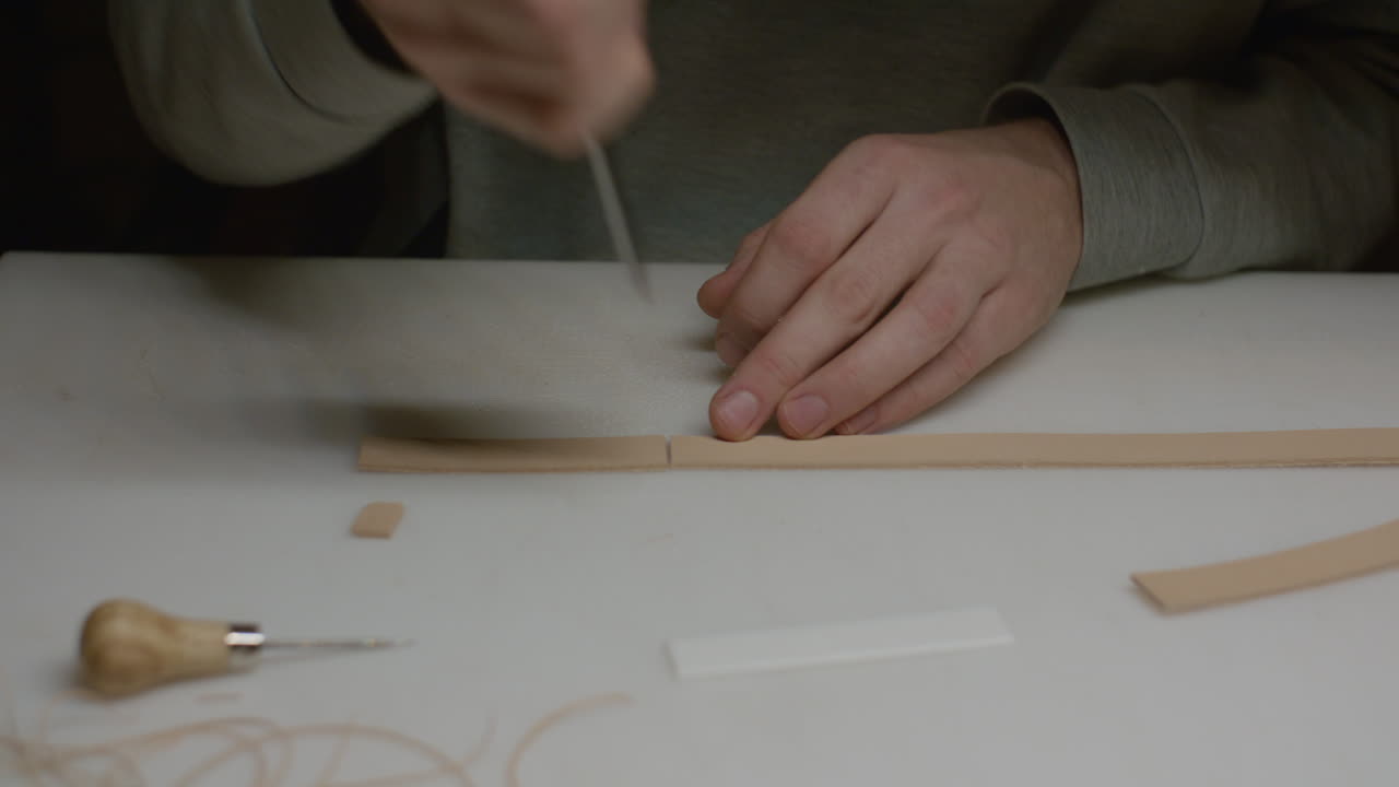 A leatherworker carefully cuts a slit into a leather strap using a sharp skiving knife on a white cutting board.