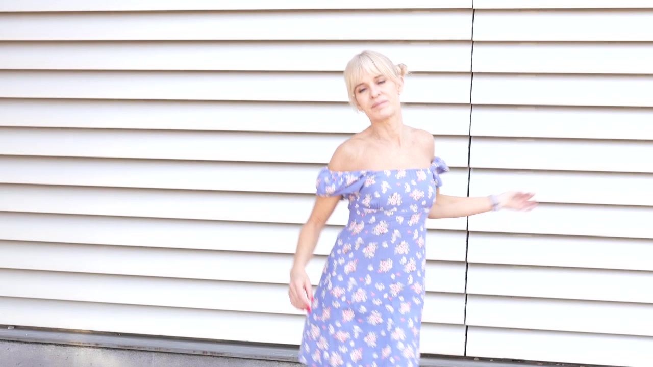 Uma mulher elegante em um vestido floral passeia ao lado de uma parede texturizada sob a luz natural do sol.