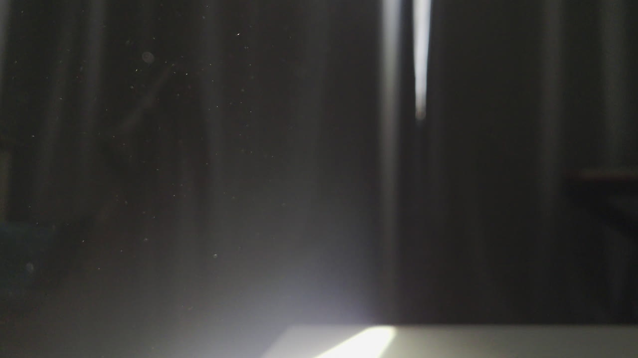 Filmagem atmosférica de alto contraste de minúsculas partículas de poeira iluminadas por um único feixe de luz filtrando por uma janela. A cena captura um movimento etéreo flutuante contra um fundo profundo e sombrio.