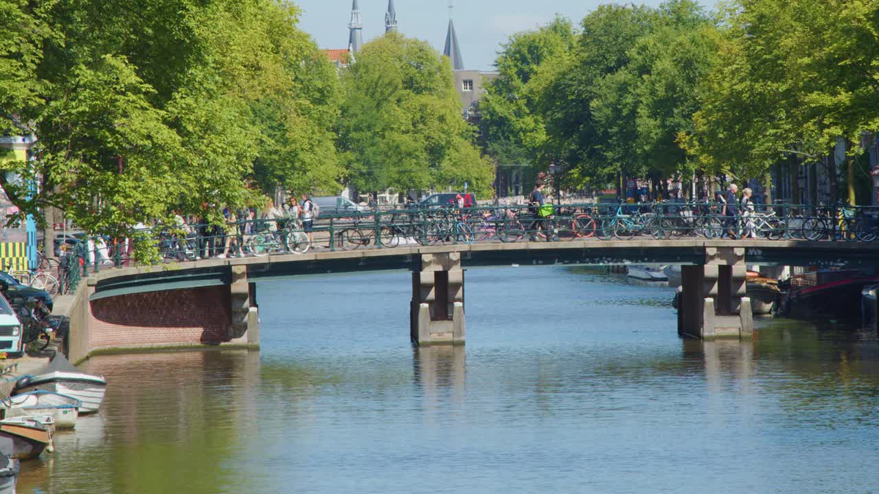 Busy Amsterdam canal bridge with cyclists, pedestrians, boats, and lush trees under bright daylight