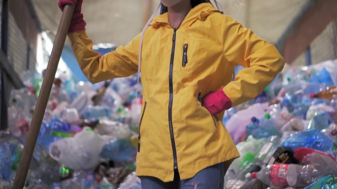 Portrait of a girl with dreadlocks, in yellow jacket and mask, holding large duck shovel, standing at a plastic recycling factory. Huge pile of bottles on background. Low angle view, turning head to the camera