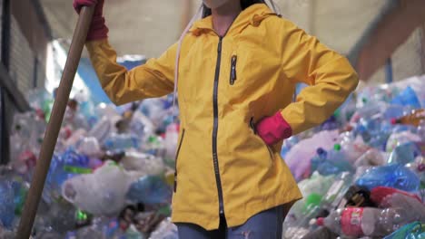 retrato de una chica con rastas, con chaqueta amarilla y máscara, sosteniendo una gran pala de pato, de pie en una fábrica de reciclaje de plástico. enorme pila de botellas en el fondo. vista de bajo ángulo, girando la cabeza hacia la cámara