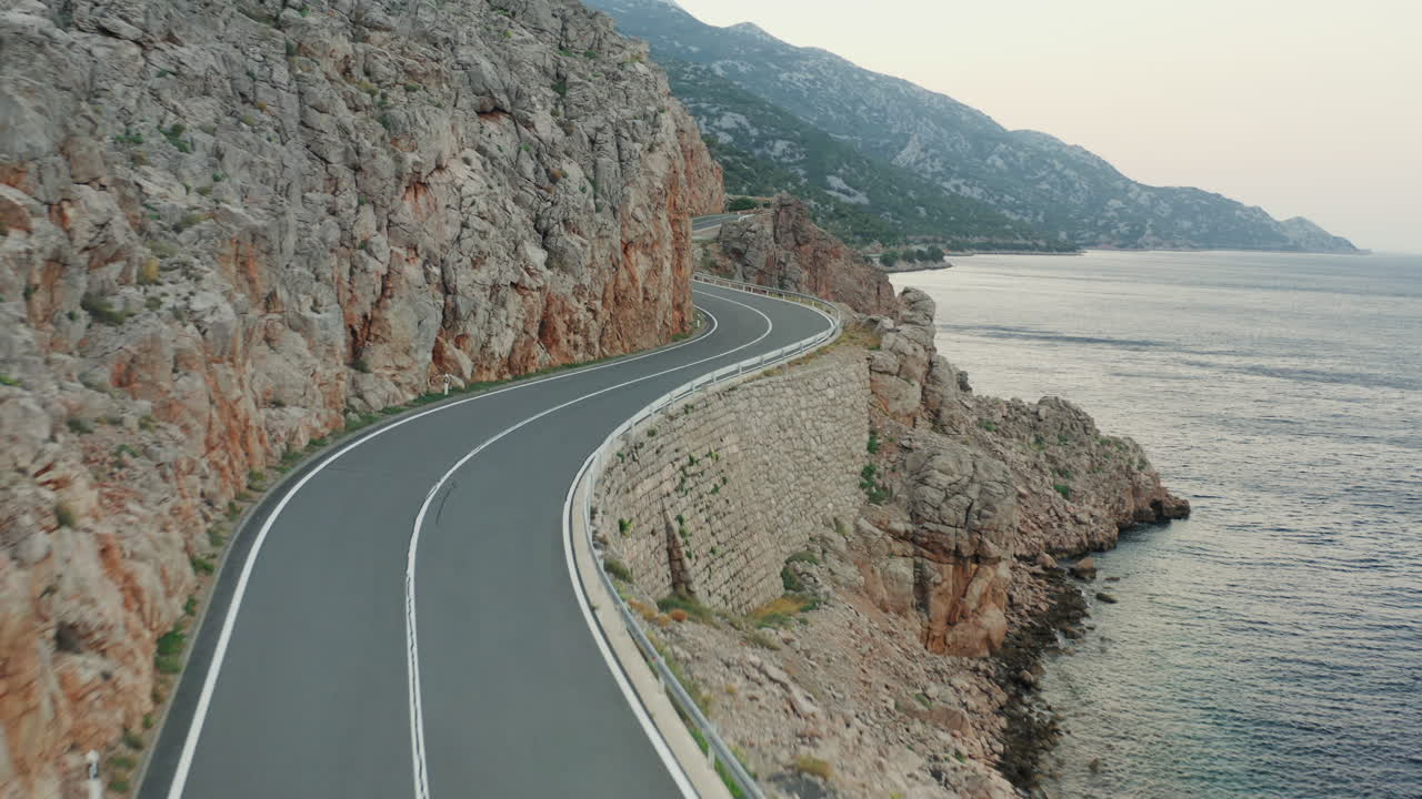 Aerial push in above winding coastal road carved into rocky cliffside at twilight. Calm sea on the right. Jadranska Magistrala, Croatia