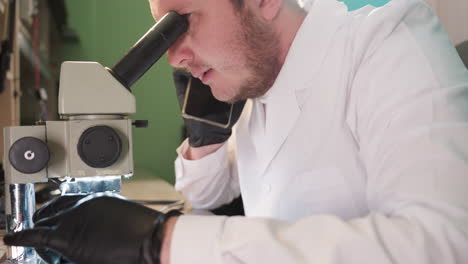 una vista de cerca de un hombre con una bata blanca de laboratorio, con guantes negros, hablando por teléfono mientras trabaja en su escritorio de laboratorio