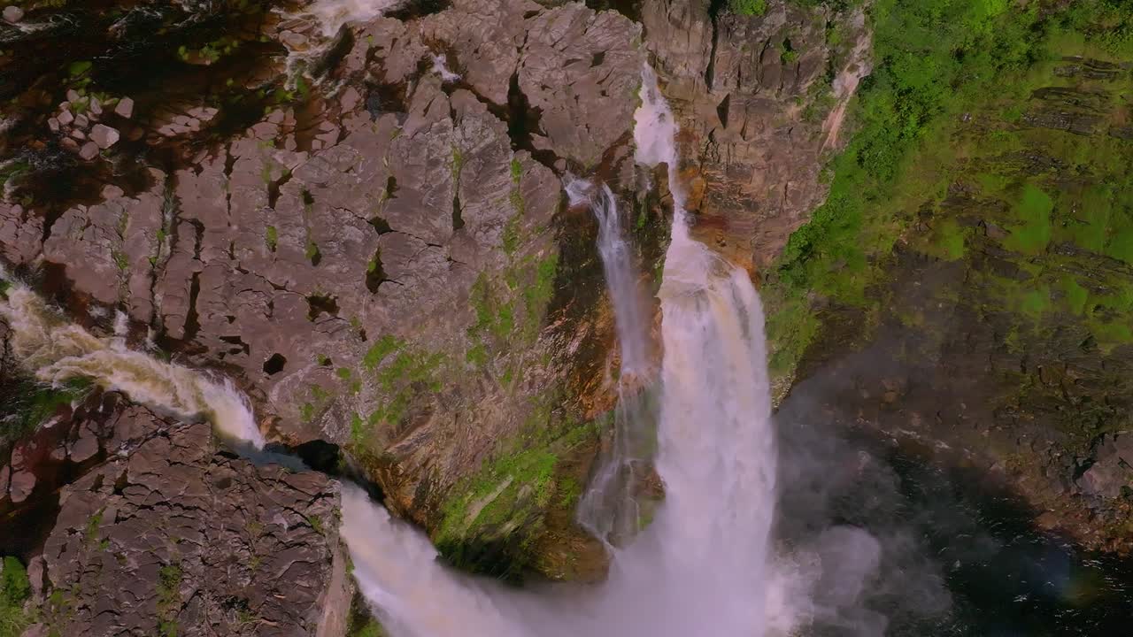 Stunning aerial view of a waterfall cascading down rocky cliffs in a lush green forest in Chapada Dos Veadeiros, Brazil, creating a rainbow, showcasing raw beauty, slow motion drone ascending shot